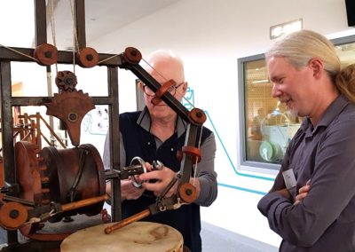 Two men standing beside da Vinci's "robot drummer" - a skeleton-like, lifesize version of a drummer robot playing a drum. The robot is made of wood, metal and cloth, and holds drumsticks.