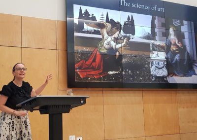 A woman at a le speaking animatedly in front of a screen featuring an artwork by Da Vinci.